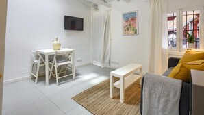 Una sala de estar con una mesa blanca, un sofá con un cojín amarillo, un televisor montado en la pared y una ventana con cortinas.