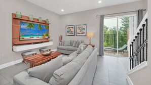 A modern living room with a sofa, a coffee table, and a flat-screen TV mounted on the wall. There are framed pictures above the sofa and a lamp on a side table.