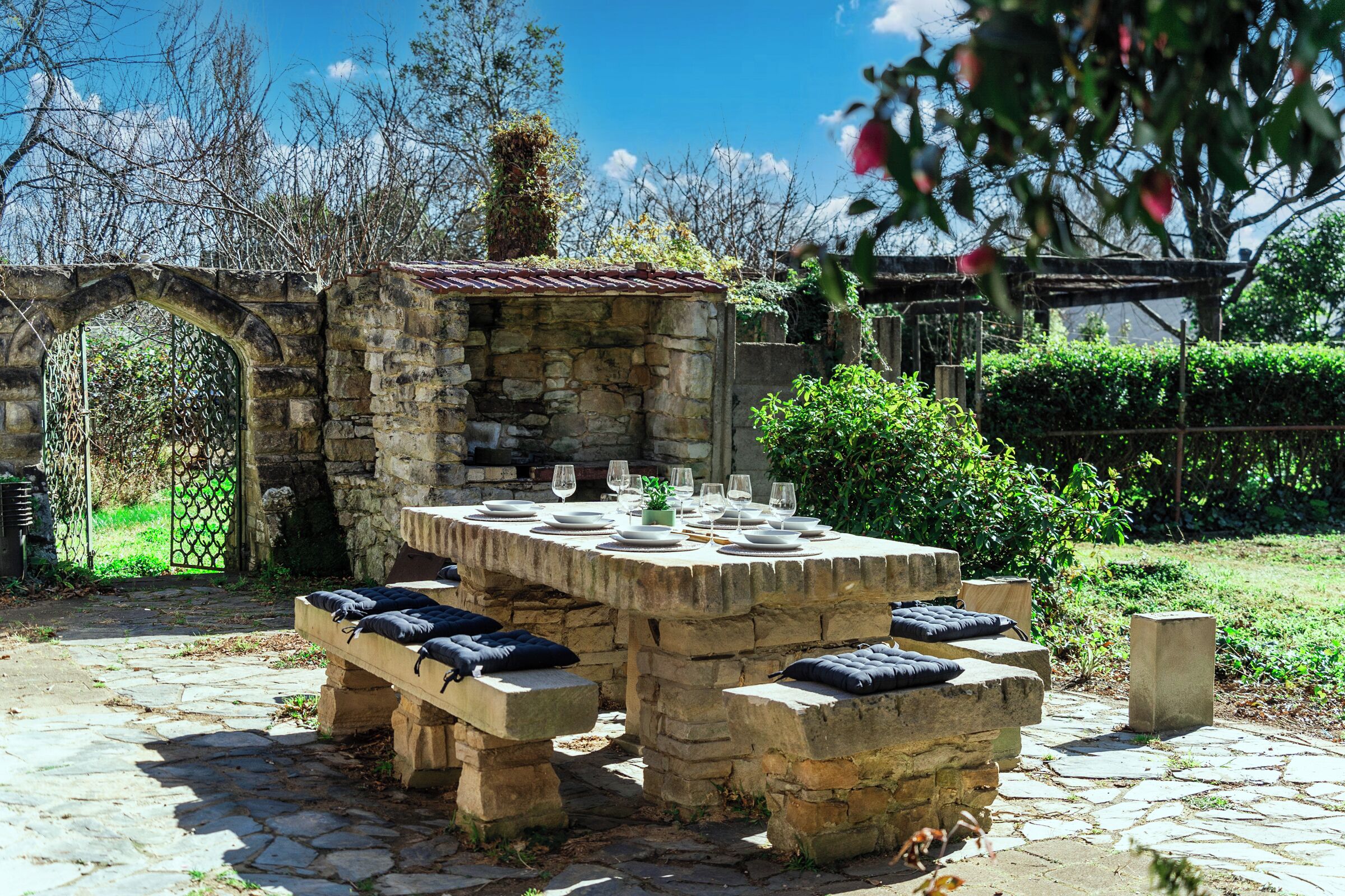 An outdoor stone table set for dining with benches, surrounded by greenery and a stone wall.