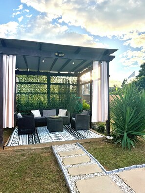 An outdoor patio area with a covered seating arrangement, a patterned rug, and a garden pathway.