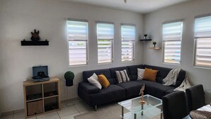 A modern living room with a dark-colored sofa, a glass coffee table, and a small side table. There are three windows with blinds, a wall-mounted shelf with decorative items, and a door with a metal gate.