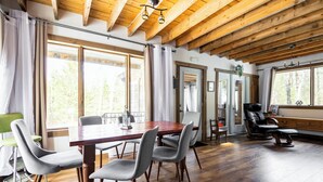 Une pièce spacieuse avec un plafond en bois, une table à manger avec des chaises, un fauteuil inclinable en cuir et de grandes fenêtres donnant sur la forêt.