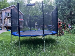 A trampoline in a grassy area with a wooden house and a swing set in the background.