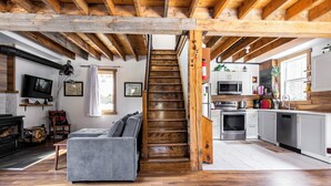 Un vaste espace de vie comprenant un canapé gris, un plancher en bois, une cheminée et une cuisine avec des armoires blanches et un escalier en bois.