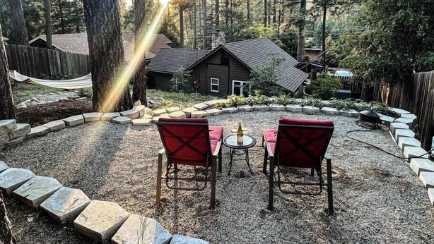A cozy outdoor seating area with two red chairs and a small table, surrounded by a stone border and a wooden fence, overlooking a forested landscape with a house in the background. - The Nest: Serene Sanctuary w Hot Tub, Fireplace, Grill, Hammock, Outdoor Fire Fe (Idyllwild-Pine Cove)