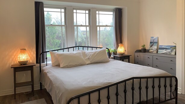 Una cama bien hecha con sábanas blancas y estructura metálica negra, flanqueada por dos mesitas de noche con lámparas, un aparador con obras de arte y vistas a la vegetación a través de la ventana.
