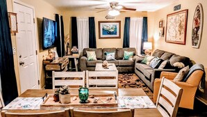 A dining area with a wooden table and chairs, a ceiling fan, and a living room in the background with a sofa and a television. - COASTAL CORAL -A few steps to the Beach, Pier, Park & close to all Boat Ramps.  (Port O'Connor)