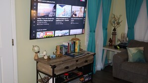 A living room with a TV mounted on the wall displaying YouTube videos, a wooden coffee table with books and decorative items, a couch, and a small side table with a plant. - COASTAL CORAL -A few steps to the Beach, Pier, Park & close to all Boat Ramps.  (Port O'Connor)