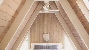 A high-ceilinged attic bedroom with wooden beams, a bed, and a hanging macrame decoration. - Clarity Cabin A-Frame Sanctuary w Hot Tub, Fireplace, Grill, AC, Hammock Chairs, Fire Pit, Pets OK (Idyllwild-Pine Cove)
