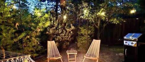 Un hamac, deux chaises, une petite table et un barbecue dans un jardin avec des arbres et une clôture.