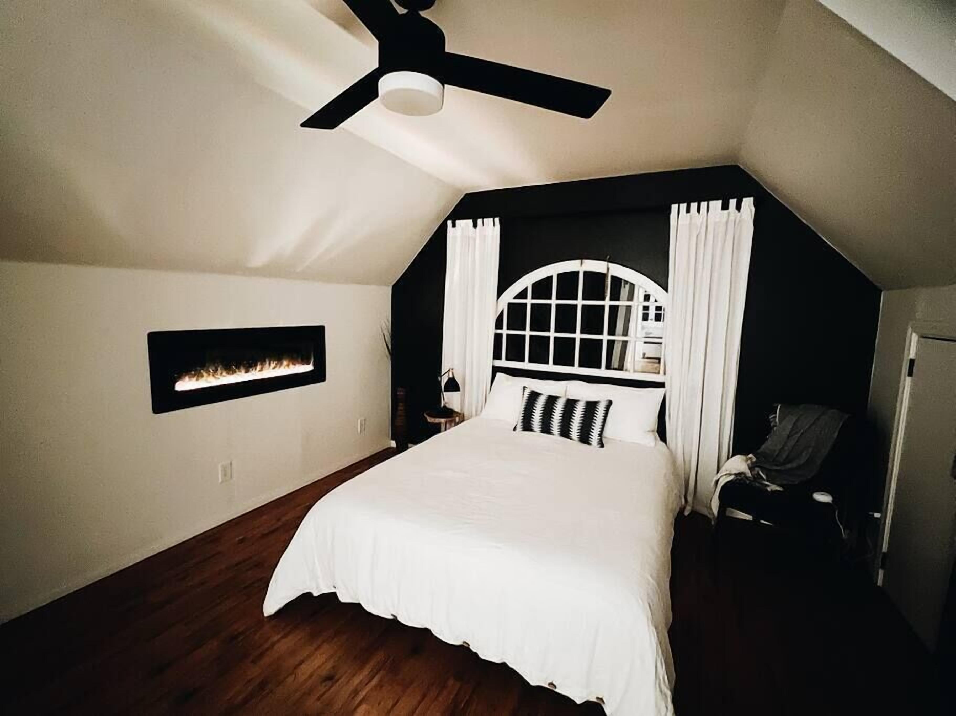 A bedroom with a sloped ceiling, a bed with white bedding, a ceiling fan, a wall-mounted fireplace, and a window with curtains.
