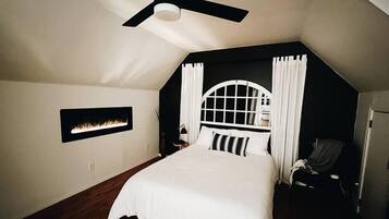 A bedroom with a sloped ceiling, a bed with white bedding, a ceiling fan, a wall-mounted fireplace, and a window with curtains.