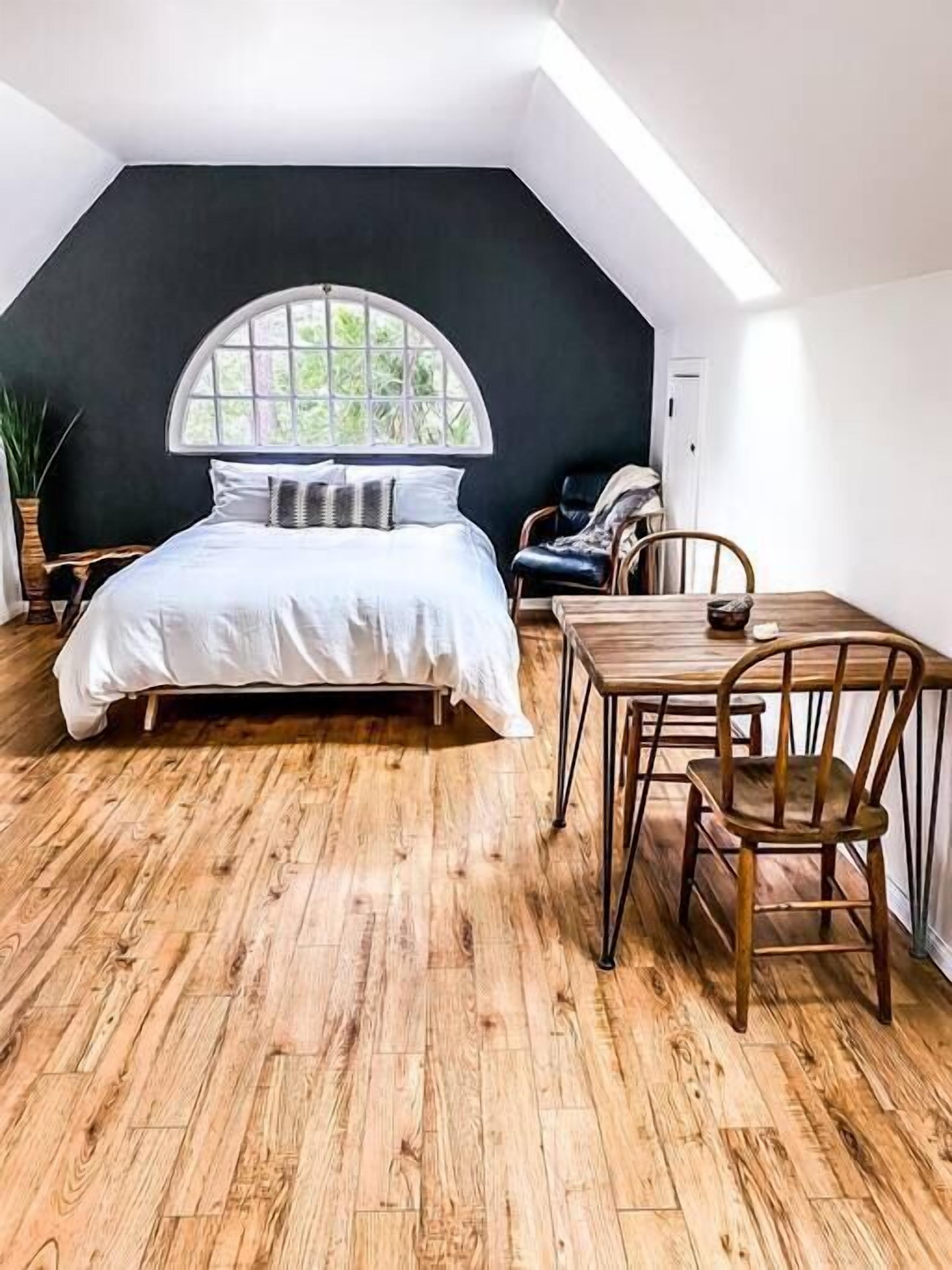 A bedroom with a bed, a dining table with chairs, a large window, and a black accent wall.