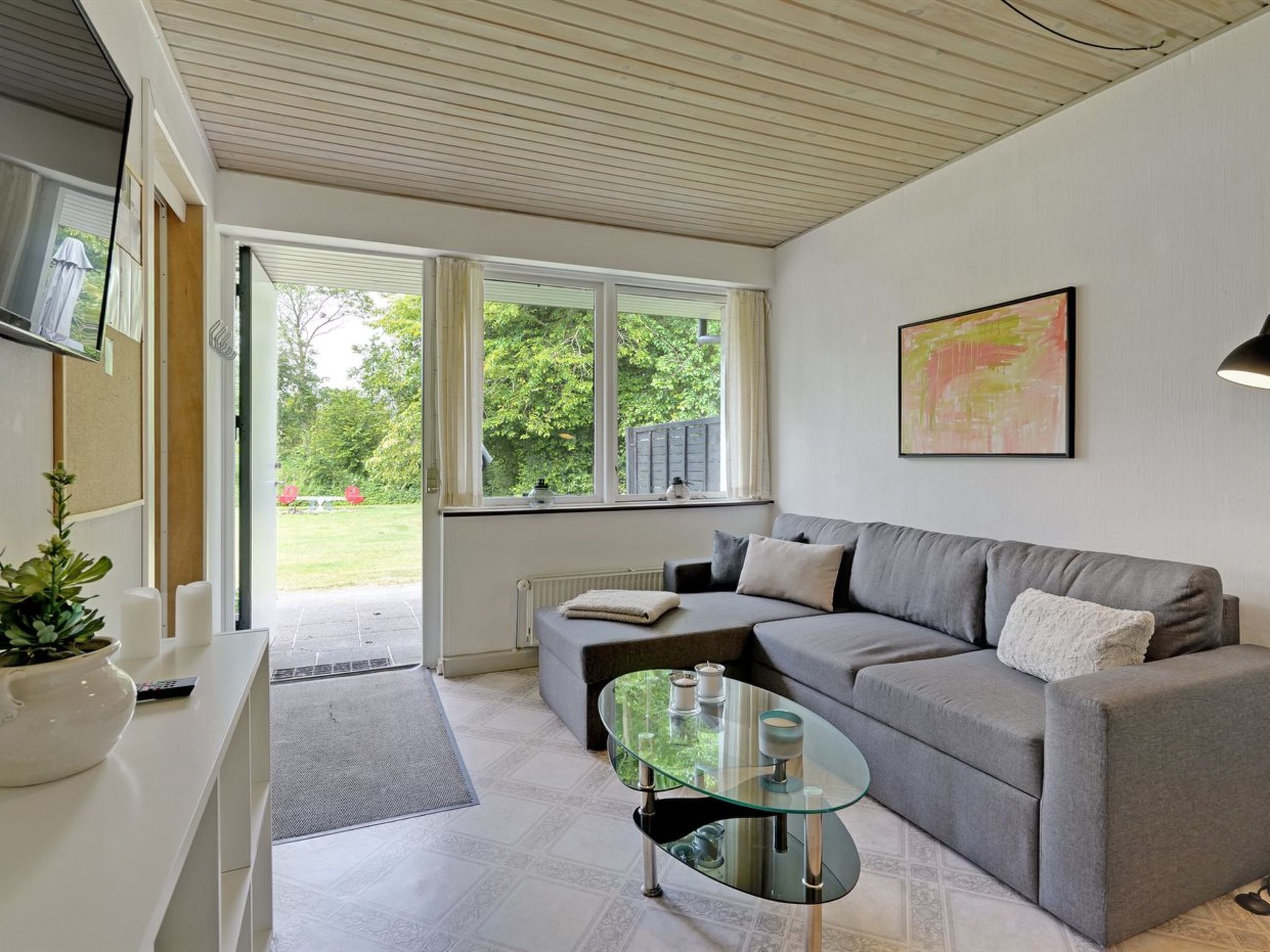 A modern living room with a grey sofa, a glass coffee table, and a view of a garden through large windows.