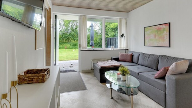 A modern living room with a grey sofa, a glass coffee table, and a large window offering a view of a garden.