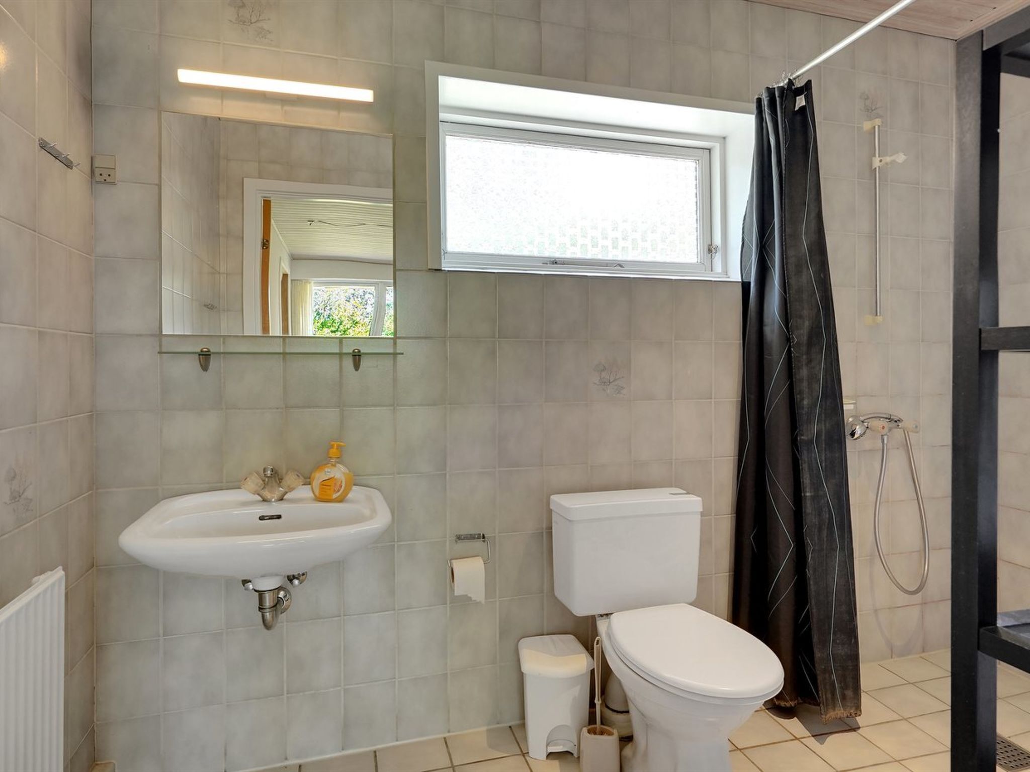 A bathroom with a white sink, toilet, and shower. There is a window above the sink and a mirror on the wall.