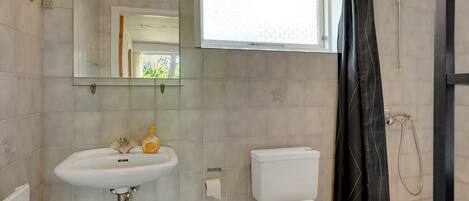 A bathroom with a white sink, toilet, and shower. There is a window above the sink and a mirror on the wall.