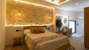 A bedroom with a stone wall, a bed with a wooden frame, a bedside table, a potted plant, and a bathroom with a shower and a mirror. - Palatina Concept Suites (Granada)