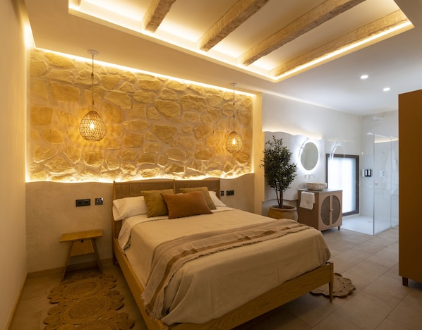 A bedroom with a stone wall, a bed with a wooden frame, a bedside table, a potted plant, and a bathroom with a shower and a mirror. - Palatina Concept Suites (Granada)