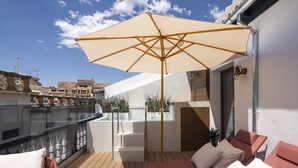 A rooftop terrace with wooden flooring, two red lounge chairs, a beige umbrella, and a view of buildings in the background. - Palatina Concept Suites (Granada)