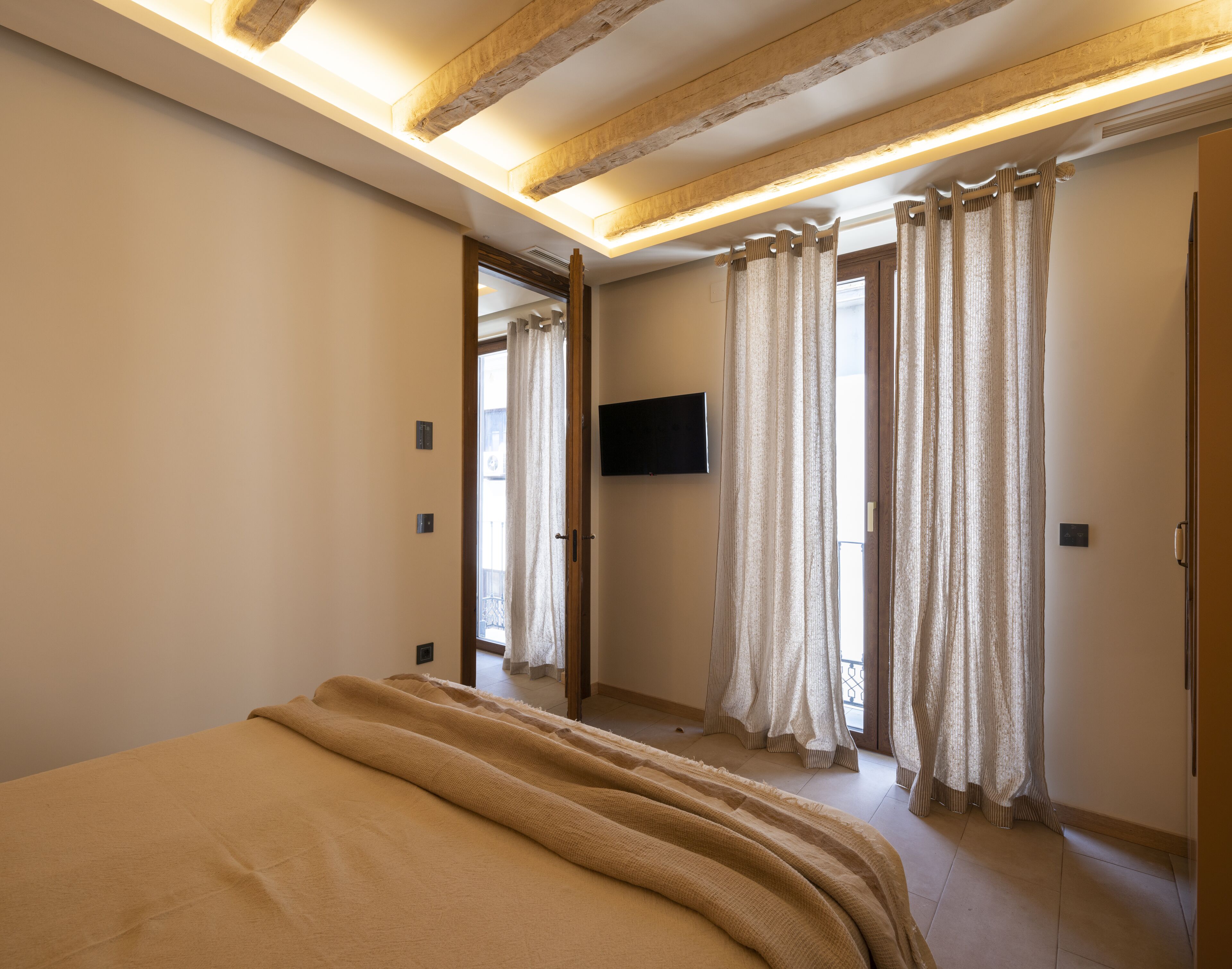 Ein Schlafzimmer mit einem großen Bett, Holzbalkendecke, einem flachbildfernseher an der Wand und einer Tür zu einem anderen Raum.