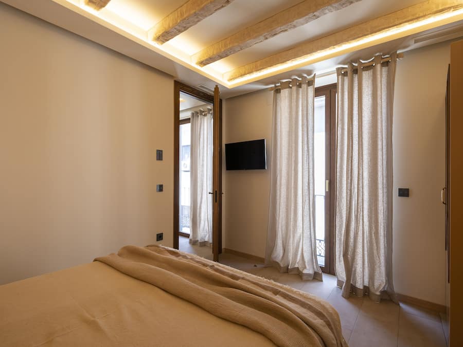 Ein Schlafzimmer mit einem großen Bett, Holzbalkendecke, einem flachbildfernseher an der Wand und einer Tür zu einem anderen Raum.
