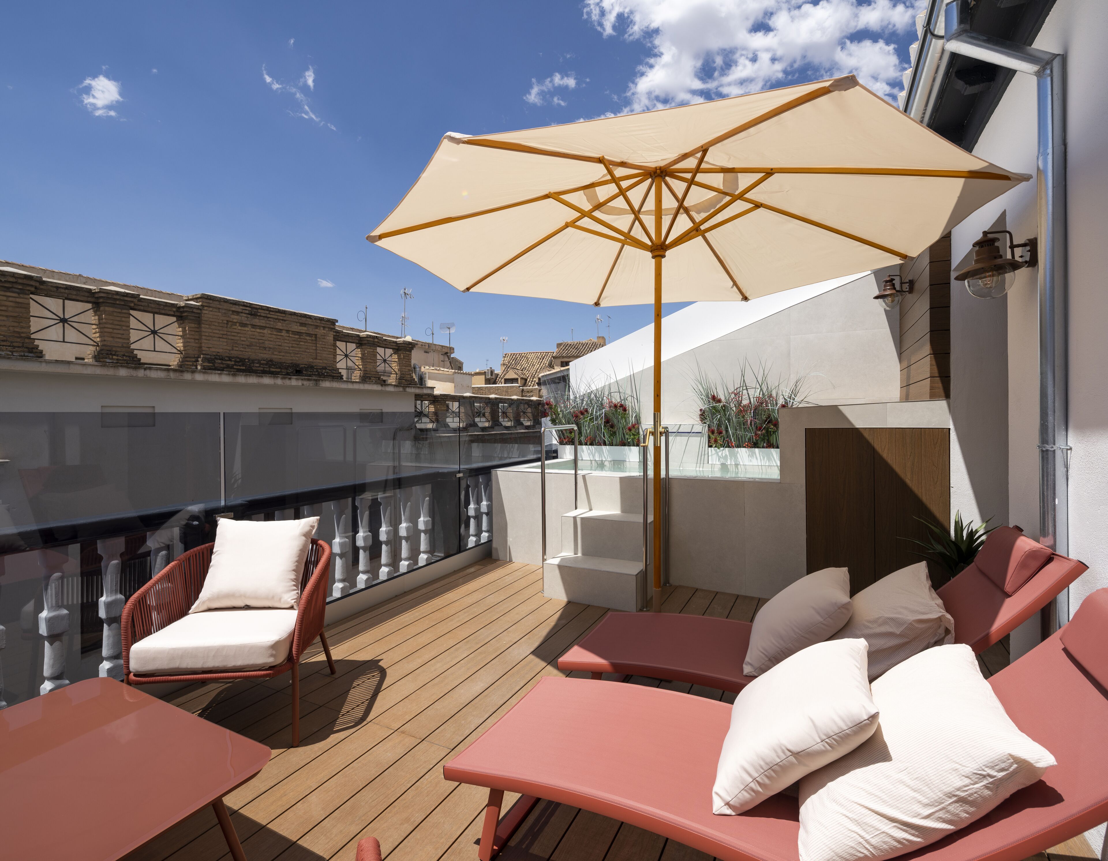 Terraza en la azotea con suelo de madera, dos sillones rojos con cojines blancos, un parasol beige y una barandilla de cristal.