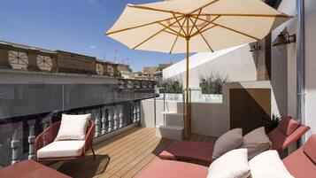 Terraza en la azotea con suelo de madera, dos sillones rojos con cojines blancos, un parasol beige y una barandilla de cristal.