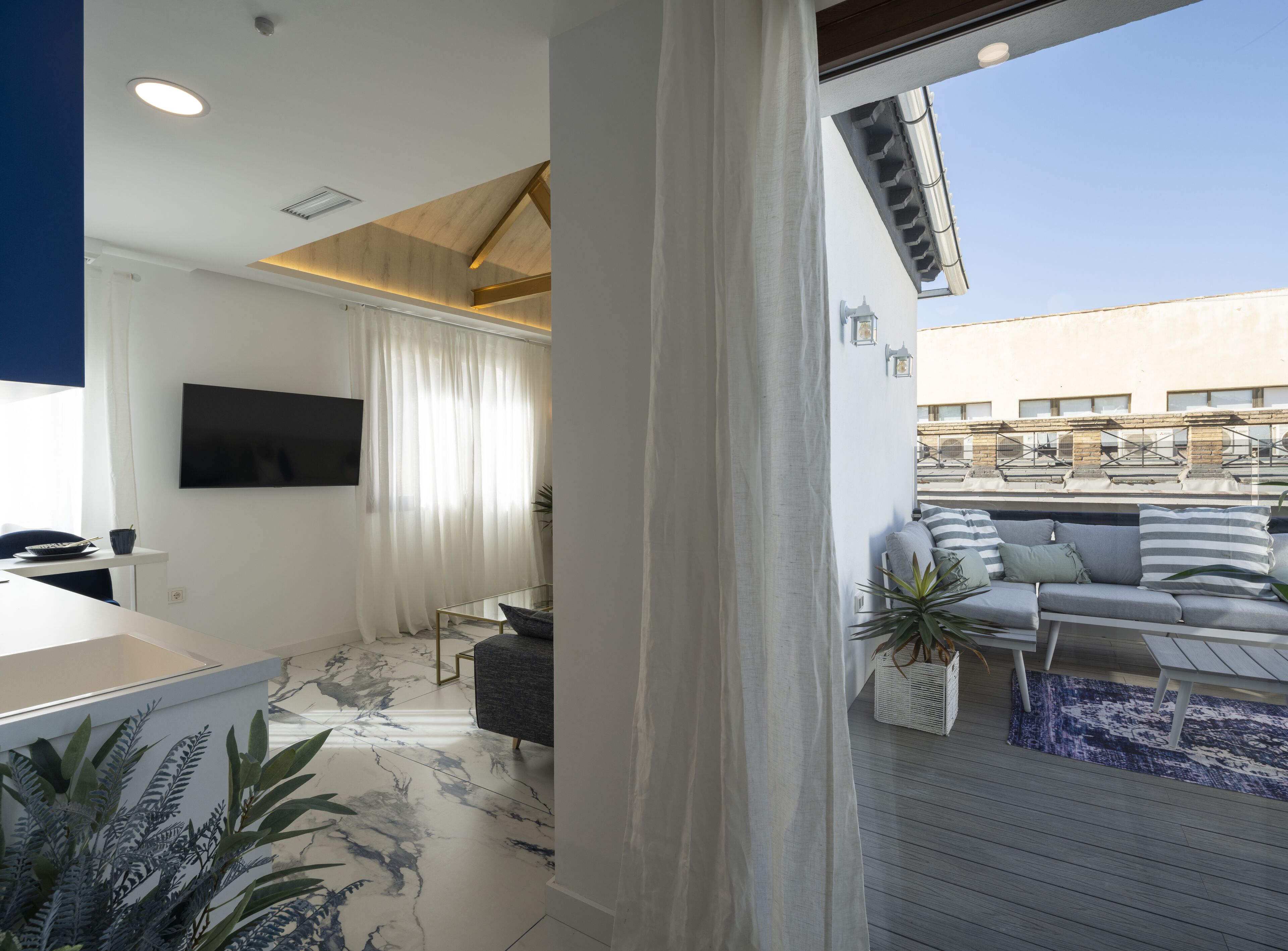 Una sala de estar moderna con un televisor de pantalla plana, una zona de comedor con mesa y sillas, y un balcón con sofá y planta en maceta.
