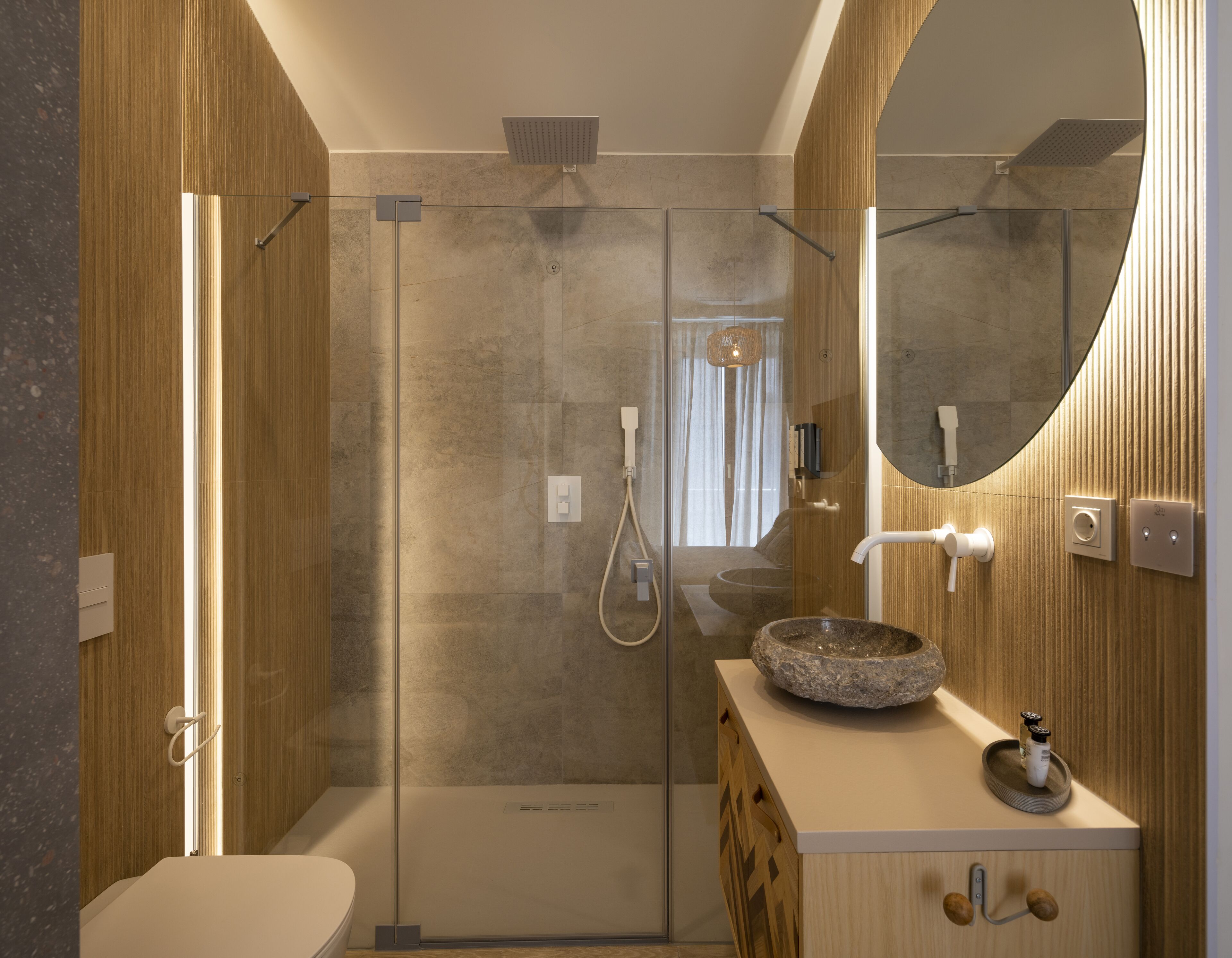 Un baño moderno con ducha envuelta en vidrio, tocador de madera con lavabo de piedra y un espejo grande.