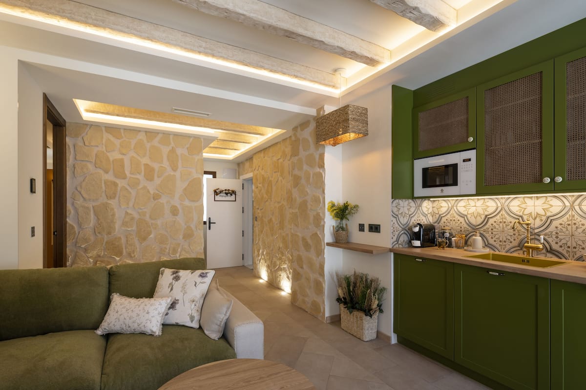 A modern kitchen with green cabinets, a microwave, and a sink. There is a stone wall and a wooden door leading to another room.