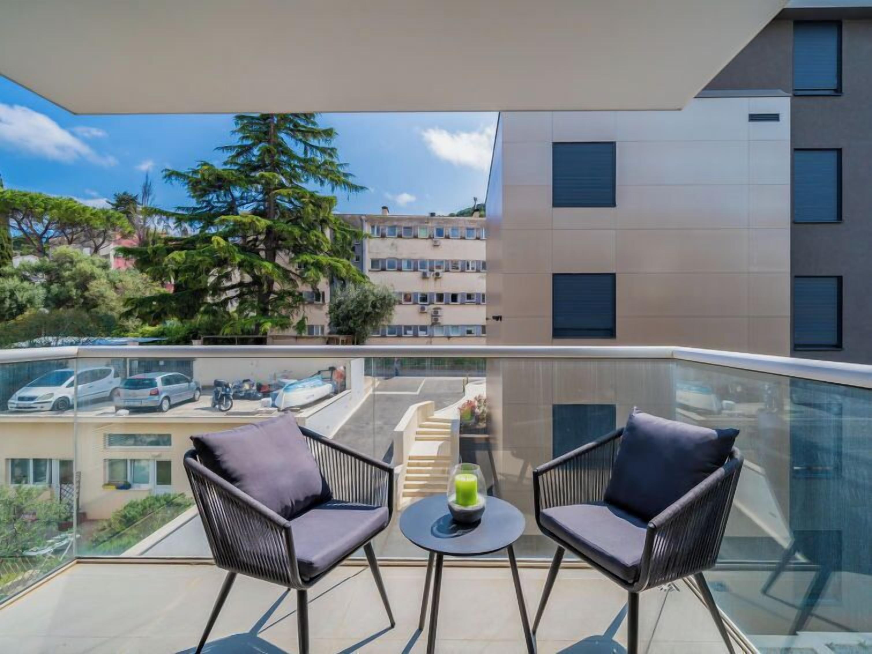 A balcony with two chairs and a small table, overlooking a parking area and a building with multiple windows.