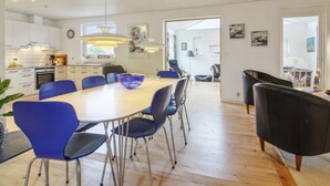 Una cocina moderna con armarios blancos, un comedor con una mesa grande y sillas azules, y un sofá negro en la sala de estar.