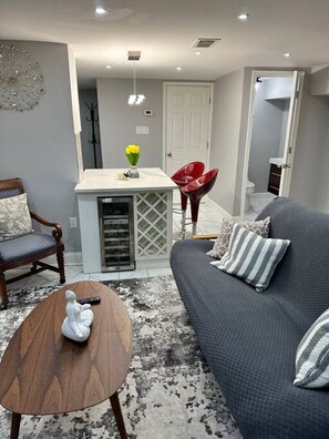 A modern living room with a grey sofa, a wooden coffee table, and a red chair. There is a small kitchen area with a white countertop and a vase with yellow flowers.
