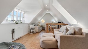 A spacious attic room with a high ceiling, wooden flooring, a sofa, a small table, and a window with plants.
