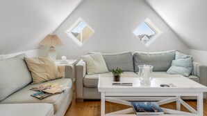 A cozy loft bedroom with a sloped ceiling, two skylights, a white sofa, a coffee table, and a bedside table with a lamp.
