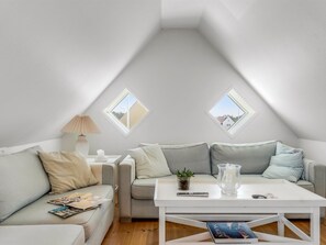 Une chambre à coucher confortable, aménagée en mezzanine, avec un plafond en pente, deux fenêtres de toit, un canapé blanc, une table basse et une table de chevet avec une lampe.