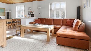 Un salon moderne avec un plafond en bois, un canapé en cuir marron, une table à manger en bois et un coin cuisine à l’arrière-plan.