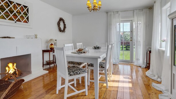 Ein Essbereich mit weißem Tisch und Stühlen, einer Chandelüster, einem Kamin und Blick ins Freie.