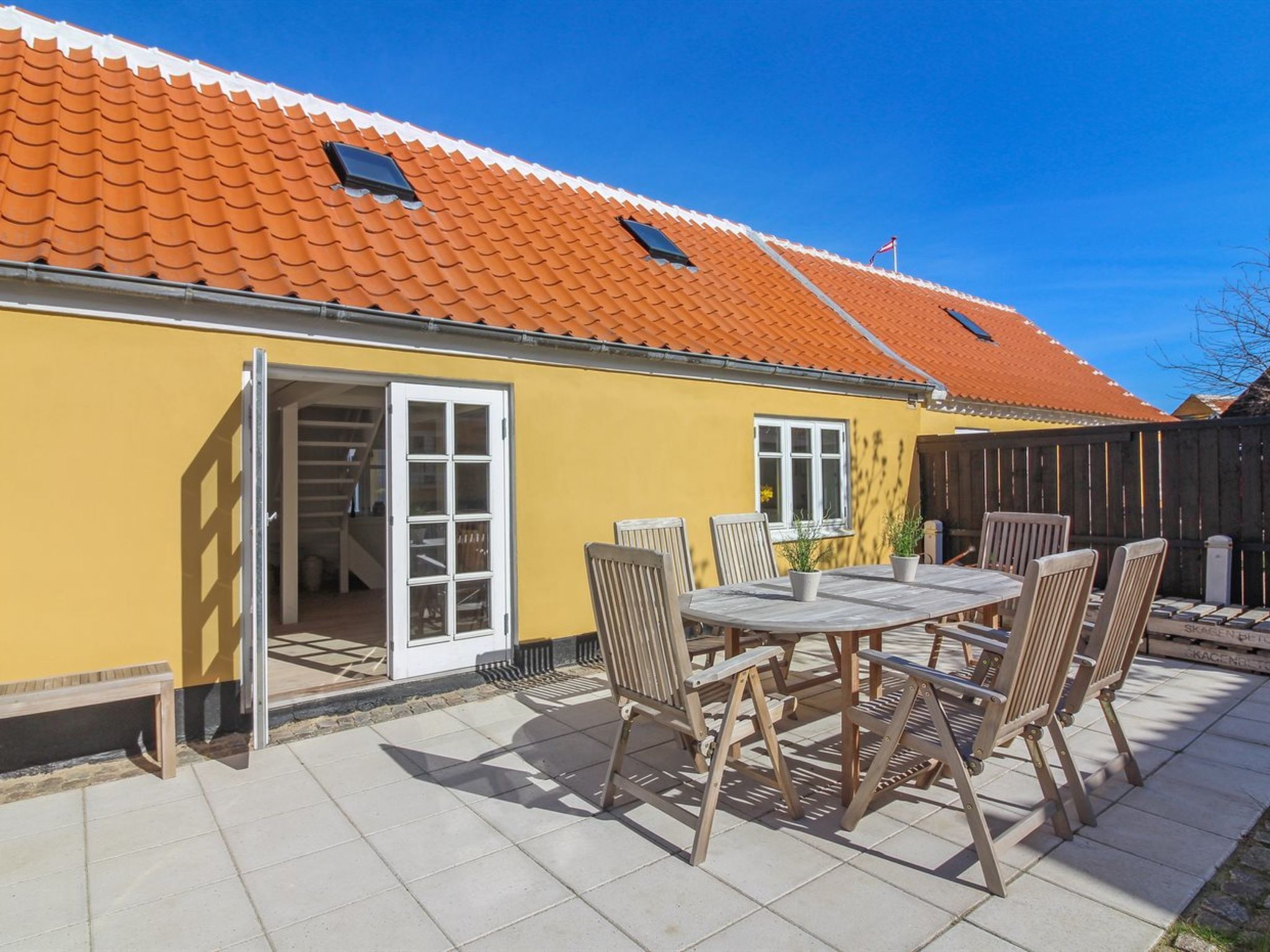 Ein gelbes Gebäude mit einem Ziegeldach, einer Terrasse mit Tisch und Stühlen und einer Tür zu einem Innenbereich.