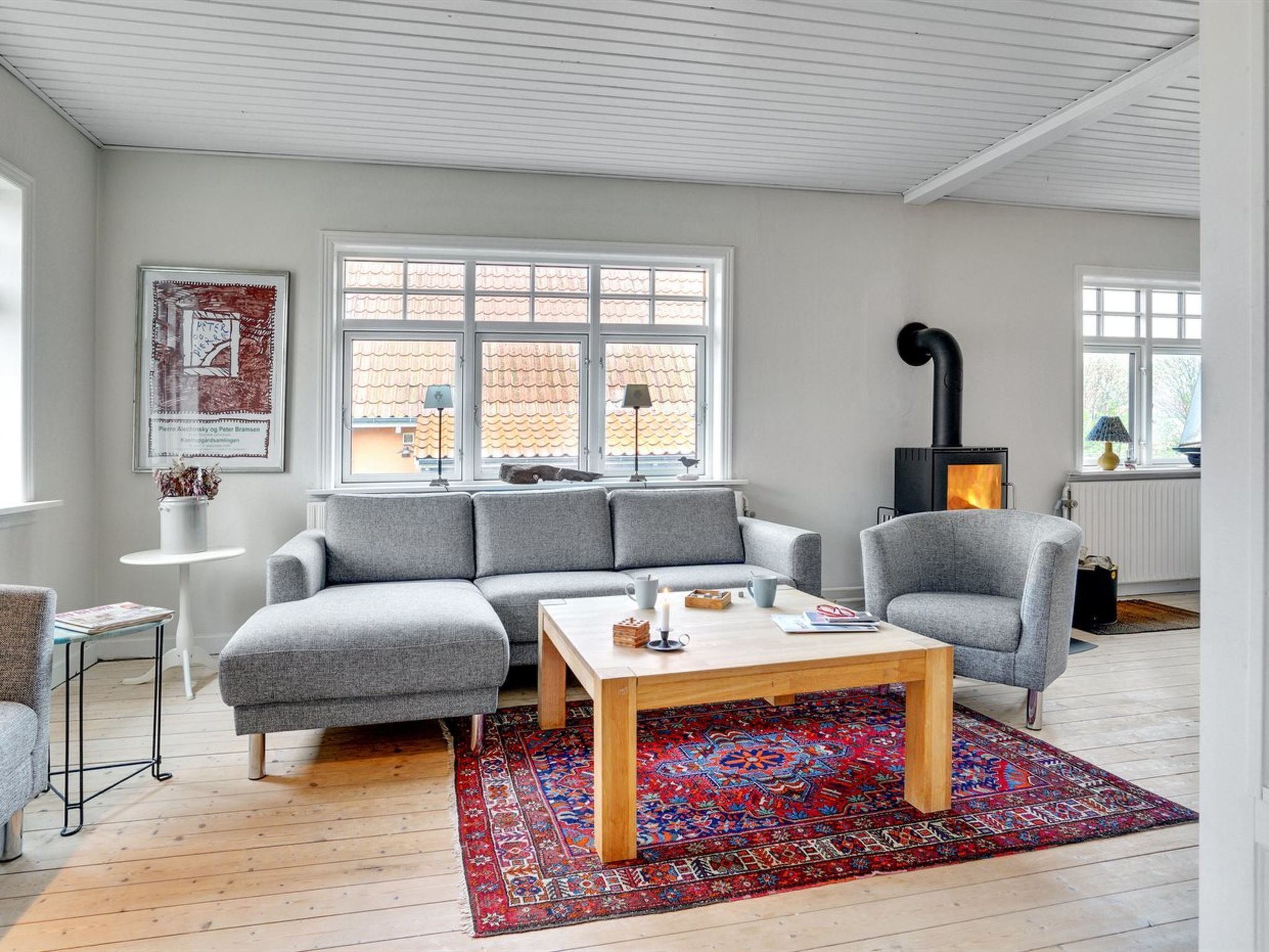 Un salon moderne avec un canapé d’angle gris, une table basse en bois et un tapis coloré. On y trouve une cheminée noire, une fenêtre donnant sur un toit recouvert de tuiles et un tableau encadré accroché au mur.