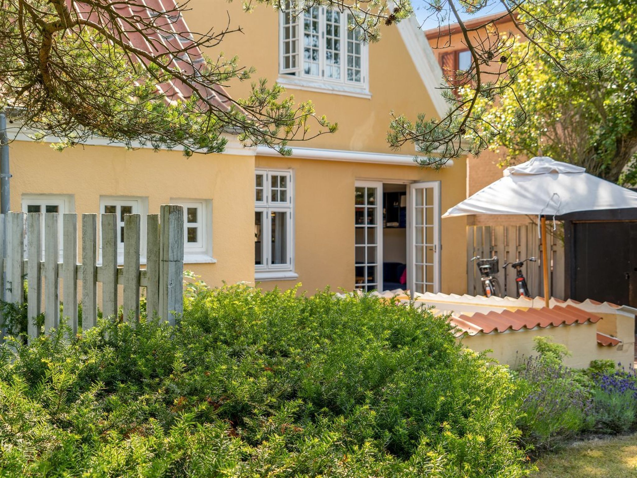 Ein zweistöckiges Haus mit gelber Fassade, einem Holzzaun und einer Terrasse mit einem weißen Sonnenschirm.