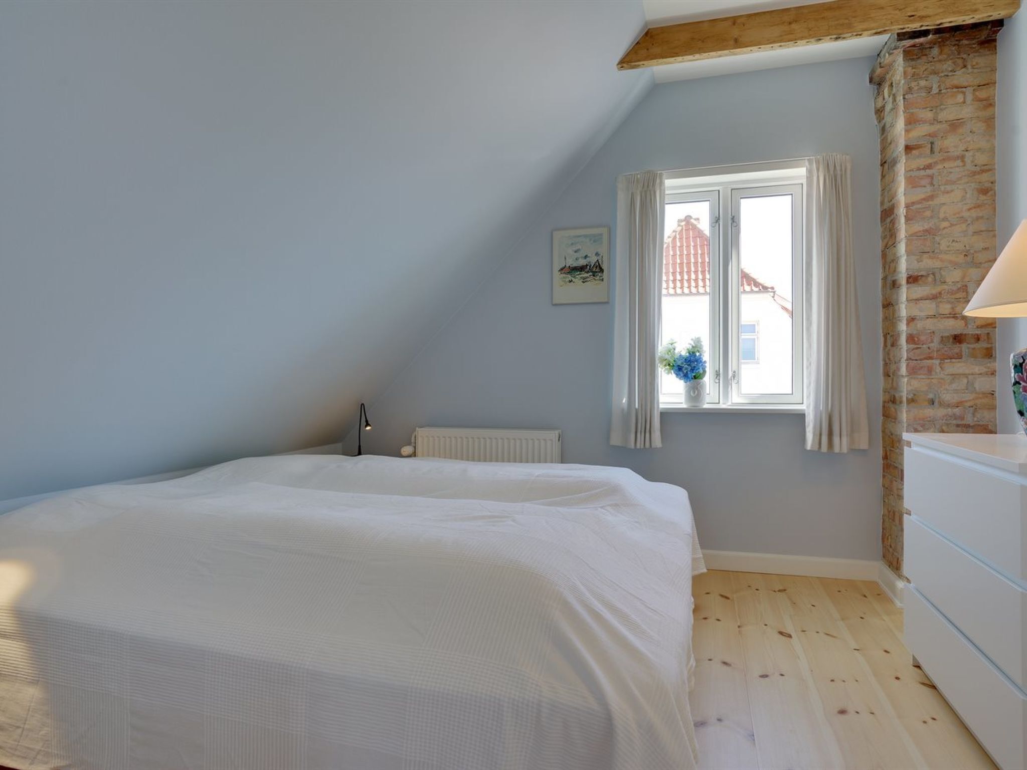 Et soveværelse med skrå vægge, en seng, en radiator, et vindue med udsigt til bygninger og en lampe.