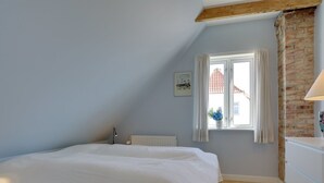 A bedroom with a sloped ceiling, a bed, a radiator, a window with a view of buildings, and a lamp.
