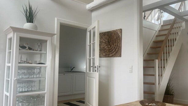 Ein Zimmer mit einem Holztisch, einer weißen Kommode mit Gläsern, einer Treppe und einem Wandbild.