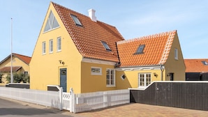 Ein gelbes Haus mit rotem Ziegeldach, weißem Zaun und einem gepflasterten Einfahrt.