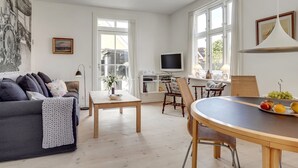 A spacious living room with a dark grey sofa, a wooden coffee table, and a dining area with a round table and wicker chairs.
