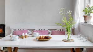 A cozy dining area with a white table, pink cushions, a white bench, a hanging light fixture, a vase with flowers, and a potted plant by the window.