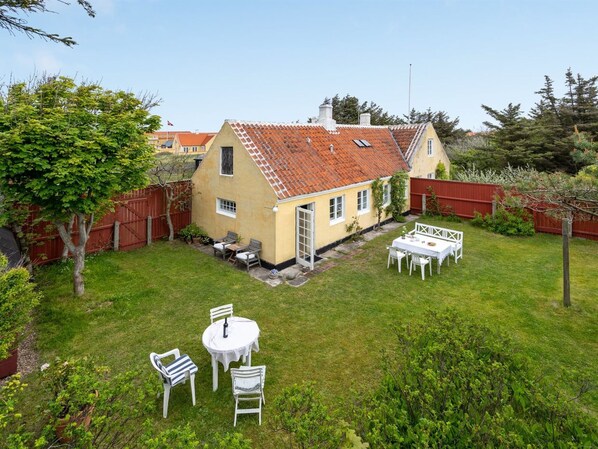 Ein Haus mit Ziegeldach, einer kleinen Terrasse mit Tisch und Stühlen sowie einem umzäunten Garten.