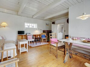 Ein gemütlicher, gut beleuchteter Raum mit Holzboden, weißen Wänden und einem Kamin. Es gibt mehrere Stühle und einen Tisch, einen Fernseher und eine Lampe.